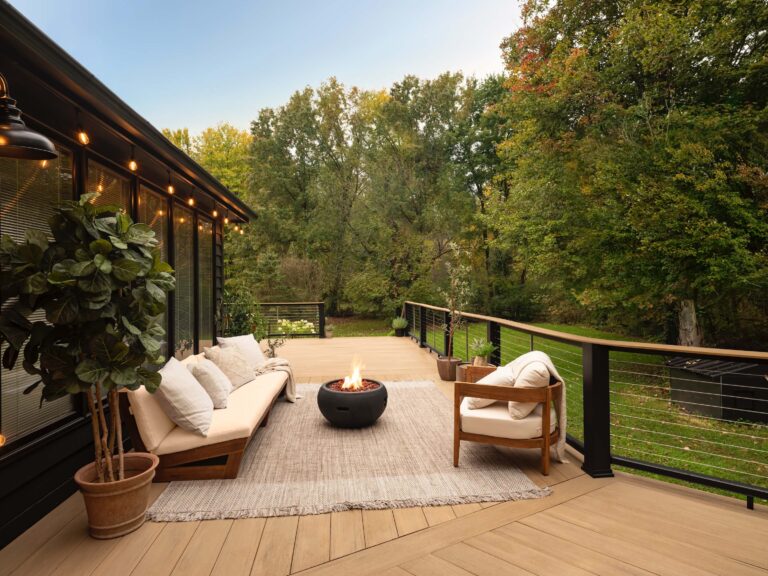 A composite deck features an expansive layout that wraps around the back of a large home. A section of the deck features an outdoor couch and matching chair sitting around a fire feature. The furniture is made of warm wood frames and cream-colored cushions for an inviting look. The decking is a sunny tan color which contrasts nicely with the black composite railing. A stainless steel cable rail infill provides clear sightlines to the lush green backyard beyond. The composite deck is made of Weathered Teak Wide-Width boards from the TimberTech AZEK Vintage Collection.