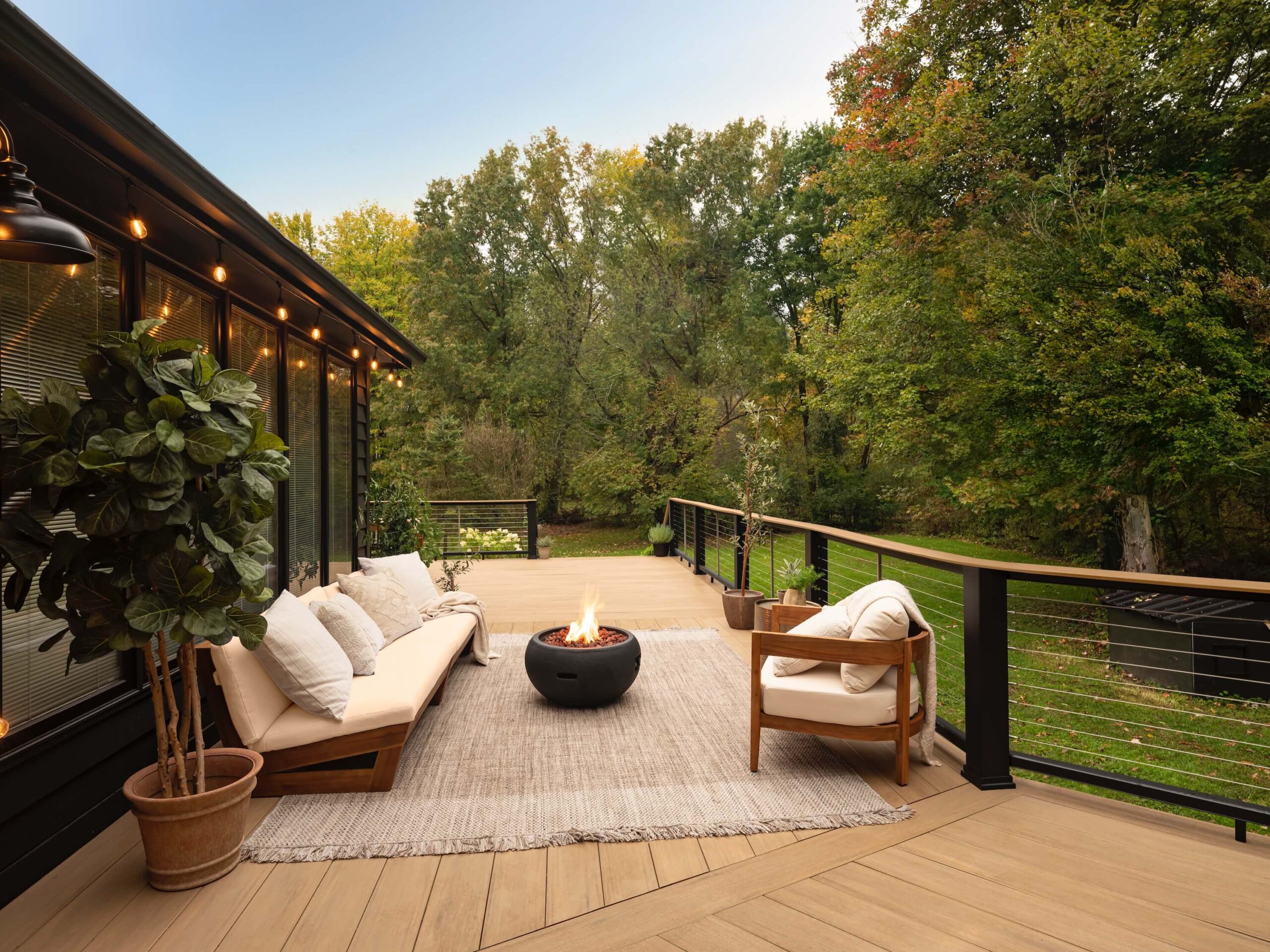 A composite deck features an expansive layout that wraps around the back of a large home. A section of the deck features an outdoor couch and matching chair sitting around a fire feature. The furniture is made of warm wood frames and cream-colored cushions for an inviting look. The decking is a sunny tan color which contrasts nicely with the black composite railing. A stainless steel cable rail infill provides clear sightlines to the lush green backyard beyond. The composite deck is made of Weathered Teak Wide-Width boards from the TimberTech AZEK Vintage Collection.